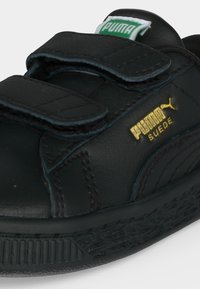 Κοντινή λήψη ενός μαύρου sneaker Puma Suede με δύο λουράκια βέλκρο και χρυσό λογότυπο στην πλευρά και πράσινη ετικέτα Puma στη γλώσσα.