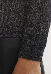 Nahaufnahme eines dunkelgrauen gestrickten Pulloverbunds, der am Handgelenk endet, über einer Hand und einem schwarzen strukturierten Stoffhintergrund.