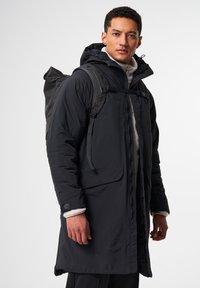 Schwarze isolierte Parkajacke mit Kapuze, Reißverschluss, Fronttaschen und strukturiertem Futter. getragen über einem leichten Pullover und einem Rucksack.