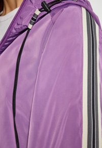 Giacca leggera viola con una superficie liscia, dotata di dettagli a righe bianche e nere lungo le maniche e un cordino regolabile.