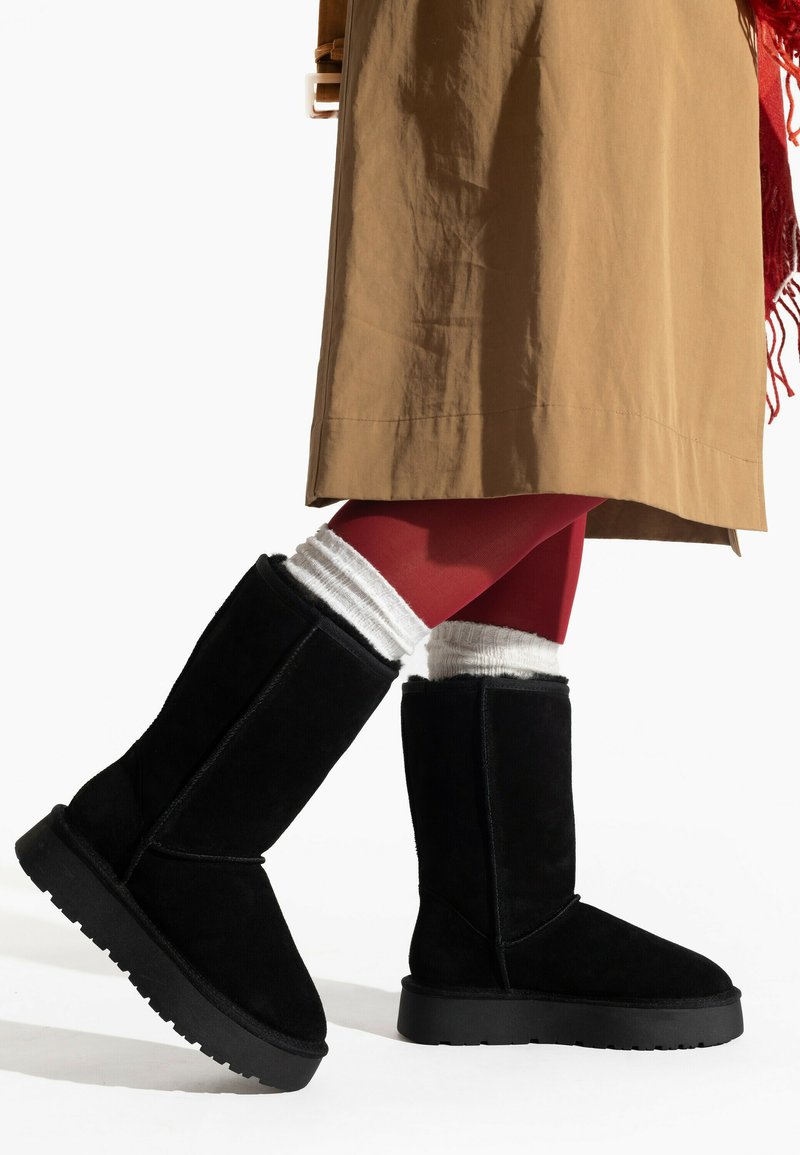 Botas altas de ante negro con superficie texturizada y suela gruesa negra, combinadas con calcetas de punto blancas y medias rojas bajo una falda color beige.