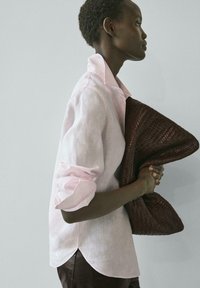 Osoba v světle růžové lněné košili drží velké tmavě hnědé pletené psaníčko, obrácená doprava na jednobarevném světlém pozadí.