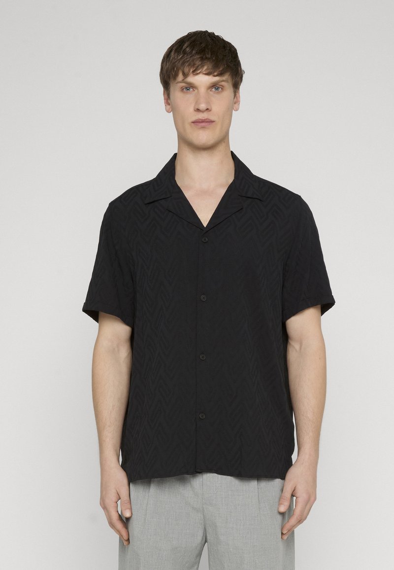 Homme portant une chemise boutonnée à manches courtes noire avec une texture en chevron subtile et un pantalon gris clair, debout devant un fond uni.