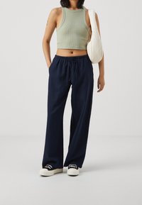 Kvinna som bär en grön crop top, marinblå breda byxor med dragsko, svart och vit sneakers, och bär en vit axelväska.