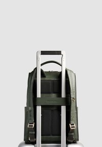 Sac à dos en cuir vert foncé fixé à la poignée d'une valise argentée, vu de dos avec les bretelles et la poignée supérieure visibles.