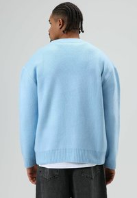 Pull oversize bleu clair avec une finition texturée, ourlet et poignets côtelés, vu de dos, associé à un pantalon foncé.