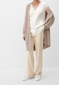 Beige kardigan přes bílou blůzu s výstřihem do V, doplněný krémovými kalhotami s širokými nohavicemi a bílými teniskami. Textura se liší mezi materiály.