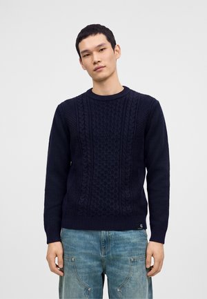 Jeune homme portant un pull torsadé bleu marine et un jean bleu clair, debout devant un fond gris uni.