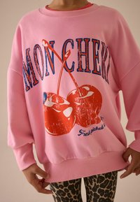 Lyserød oversized sweatshirt med blå og røde kirsebærgrafik og teksten "MON CHERI". Har ribbede manchetter og kant, båret over leopardmønstrede leggings.