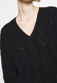 Persona che indossa un maglione nero a V in maglia con un sottile motivo a foglia, che mostra il collo e le spalle superiori su uno sfondo semplice.