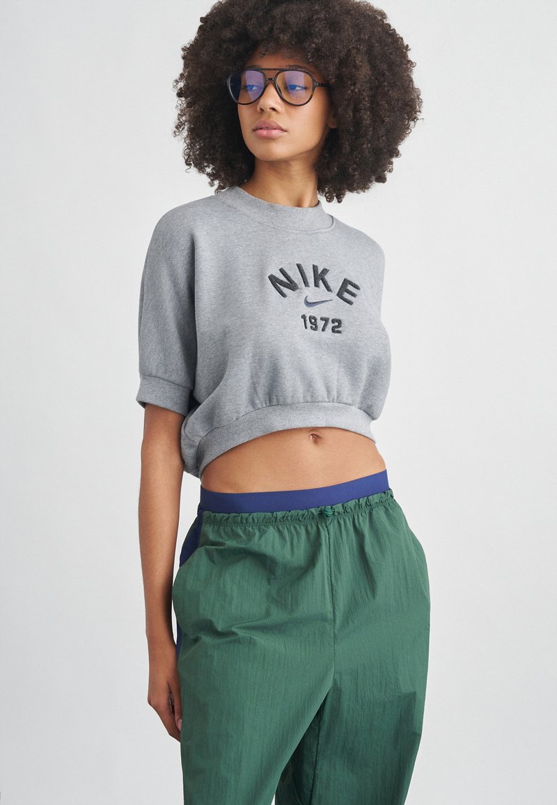 Γκρι κολλημένο φούτερ με εκτύπωση «NIKE 1972»· συνδυάζεται με πράσινο υφασμάτινο φαρδύ παντελόνι με μπλε ζώνη στη μέση και πλαϊνές τσέπες.