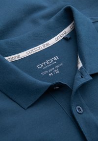 Blå polo-skjorte krave med et mærkat, der viser "ombre cotton line", 100% ren bomuld, størrelse M EU 48 og mærket inderhalsbånd.