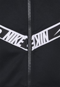 Svart zip-up sweatshirt med vita logostriper och en texturerad tyg. Dragkedjan går centralt, med vinklad färgblockering på axlarna.