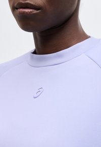 Κοντινό πλάνο ατόμου που φοράει φωτεινό μωβ μπλουζάκι με στρογγυλή λαιμόκοψη και μικρό ανάγλυφο λογότυπο Asics στο στήθος.