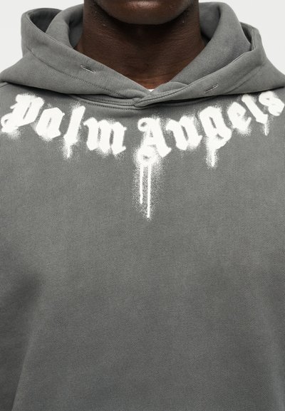 Γκρι φούτερ με κουκούλα, με έντονη λευκή γραφή που στάζει, "Palm Angels" στο στήθος. Το ύφασμα έχει απαλή υφή με casual σχεδιασμό.