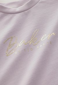 Fialová mikina s hladkým povrchem, na které se nachází velké kovově zlaté kurzívou psané logo "Baker" a text "by Ted Baker" pod ním.