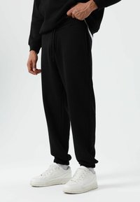 Zwarte joggingbroek met elastische boorden, een taille met trekkoord en zijzakken, gecombineerd met witte sneakers op een effen achtergrond.