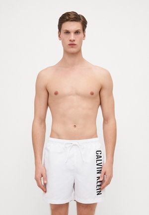 Șorturi de baie albe cu talie elastică și șnur de reglare, prezentând un logo vertical negru "CALVIN KLEIN" pe partea stângă.