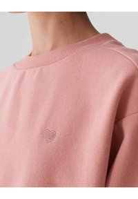 Felpa rosa con colletto a costine e piccolo ricamo a forma di cuore. Tessuto morbido, vestibilità leggermente oversize, design minimale, senza chiusure visibili.