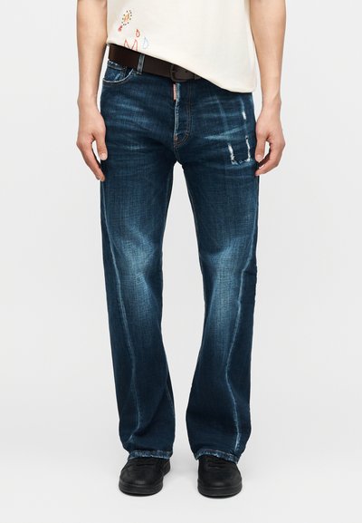 Άνδρας που φοράει μωβ-μπλε ξεθωριασμένα τζιν με μαύρη ζώνη, συνδυασμένα με μαύρα παπούτσια και λευκό πουκάμισο μερικώς ορατό.