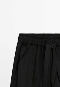 Pantalon noir avec une taille élastique et une ceinture en tissu nouée, montrant la partie avant supérieure et les poches latérales.