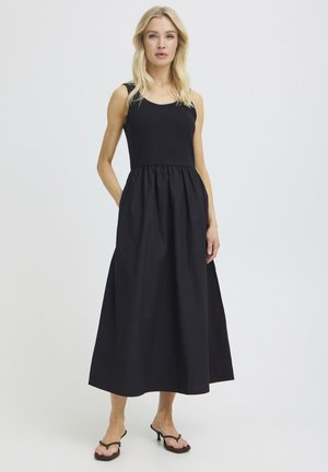 Blond naine kannab musta varrukateta maksikleiti, millel on kogutud talje, seisab ühe käega taskus tavalise tausta ees.