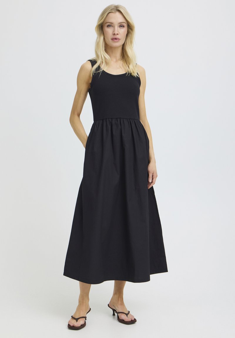 Femme blonde portant une robe longue noire sans manches avec une taille froncée, debout avec une main dans la poche sur un fond uni.
