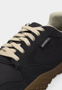 Μαύρο αθλητικό παπούτσι Merrell με μπεζ κορδόνια και υφασμένη καφέ σόλα, με την ετικέτα της μάρκας στο πλάι κοντά στα κορδόνια.