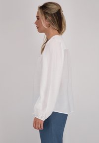 Witte blouse met lange, opgezette mouwen en gestructureerde stof. Een ronde halslijn en een iets losse pasvorm, gedragen over blauwe broeken.