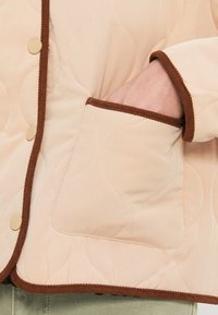 Μπεζ καπιτονέ μπουφάν με καφέ διακόσμηση, κουμπιά με πατήματα και ανοιχτή τσέπη με διακριτικό κυματιστό σχέδιο στην υφή του υφάσματος.