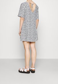 Vestido de manga corta y espalda en V con patrones de corazones negros sobre un fondo blanco. Se lleva con sandalias blancas de tiras y una superficie texturizada.
