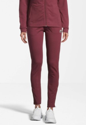 Giacca sportiva bordeaux con pantaloni aderenti abbinati, realizzati in materiale morbido. I pantaloni presentano un piccolo dettaglio con logo; le sneaker sono bianche con accenti grigi.