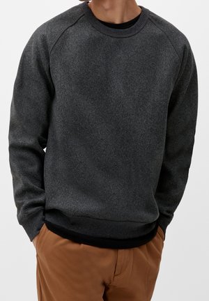 Mann trägt einen dunkelgrauen Pullover mit langen Ärmeln und braune Hose, Hände in den Taschen, vor hellem einfarbigem Hintergrund.