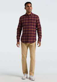 Rote und marineblaue Karobluse mit Knöpfen, kombiniert mit khaki Hosen und weißen Sneakern. Die Bluse hat lange Ärmel und einen Kragen.