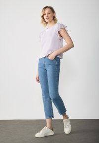 Top scurt de culoare violet deschis cu margini volan, asortat cu blugi albaștri și adidași albi, stând pe un podea gri.