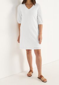 Biała sukienka z krótkim rękawkiem i dekoltem w kształcie litery V, ozdobiona wzorami z ażurowym motywem i luźnym krojem, zestawiona z brązowymi klapkami.