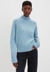 Maglione lavorato a maglia azzurro chiaro con collo alto, orlo e polsini a coste. Presenta spalle scese e una finitura texture. Abbinato a jeans neri.