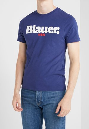 T-shirt en coton bleu marine avec un grand logo blanc "Blauer." avec des accents rouges, col rond et manches courtes, porté avec un jean bleu.