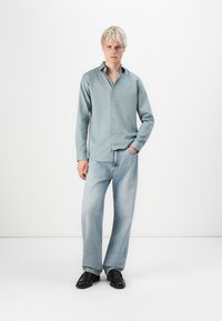 Camisa abotonada de color azul claro con mangas largas, combinada con jeans de mezclilla de lavado claro y suéteres negros. Diseño simple y moderno.