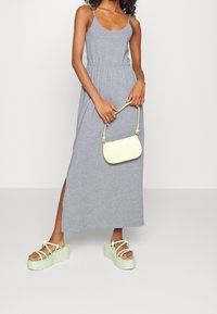 Graues ärmelloses Maxikleid mit elastischem Bund, begleitet von einer kleinen gelben Handtasche und grünen Plateau-Sandalen mit floralen Akzenten.