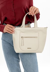 Bolso tote blanco texturizado con dos asas superiores, bolsillo frontal con cremallera y marca en dorado. Mujer sosteniendo el bolso mientras lleva una camisa burdeos.
