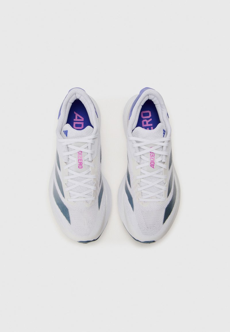 Zapatillas deportivas blancas con parte superior de malla, acentos grises y plantillas moradas. Presentan cordones, punta redondeada y una suela texturizada.