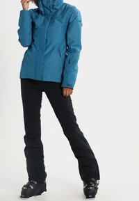 Türkisfarbene wasserdichte Jacke mit tailliertem Design, Reißverschluss-Taschen und Kapuze, kombiniert mit schwarzen Schneehosen und schwarzen Skischuhen.