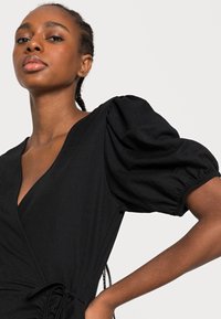 Vestido preto envolvente com mangas bufantes, um decote em V profundo e uma cintura ajustada. O tecido parece macio com uma textura suave.