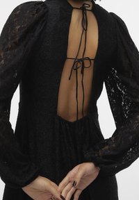 Svart spetsklänning med lång puffärm, öppen rygg som hålls på plats med en knytning och med texturerat mönster. Utsvängd kjol med rynkdetalj vid midjan.