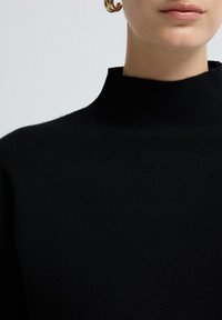 Zbliżenie na osobę w czarnym swetrze z wysokim kołnierzem i złotą kolczykiem w kształcie obręczy, ukazujące usta i dolną część twarzy.