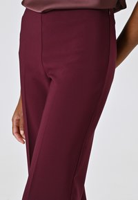 Pantalon sur mesure bordeaux avec une texture lisse, taille haute et coupe droite. Il comporte une fermeture éclair latérale et des coutures discrètes.