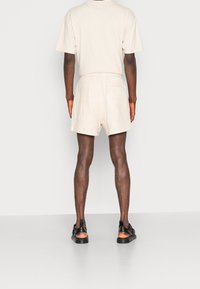 Personne debout, vue de dos, portant un short beige, un t-shirt beige et des sandales noires à bout ouvert, sur un fond clair uni.