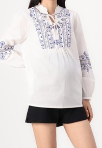Biała bluzka z niebieskimi haftowanymi kwiatami na piersi i rękawach, z okrągłym dekoltem oraz wiązaniem, w luźnym fasonie i długimi rękawami.