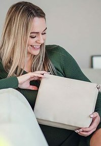 Béžová kožená taška s hladkým povrchem a minimalistickým designem, se zipovým uzávěrem a decentním logem. Drží ji osoba v zeleném svetr.